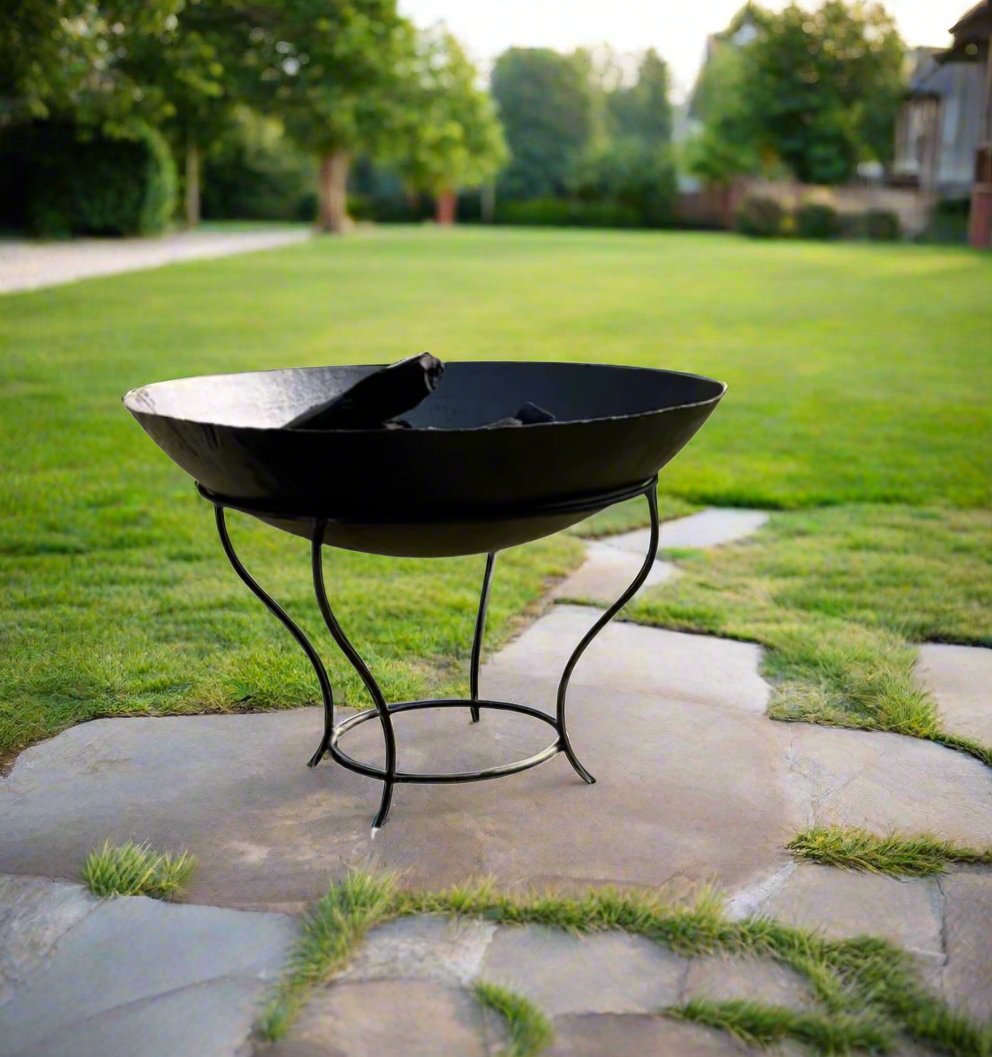 An outdoor wood and coal burning stove, known as a fire pit, placed on a stone surface with grass in the background.