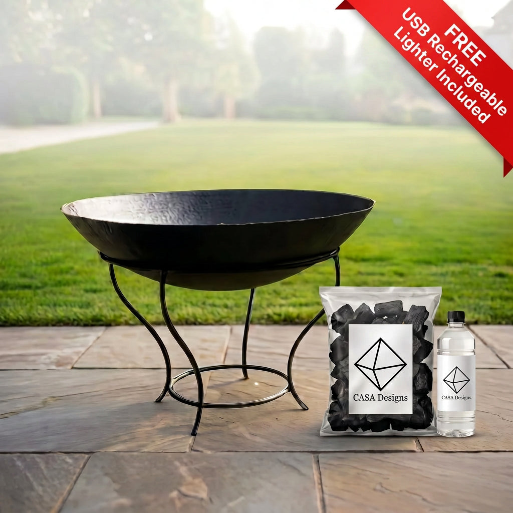 A black metal fire pit bowl on a matching stand sits on a stone patio with a blurred green lawn in the background. A bag of black charcoal and a clear bottle with a black cap are placed beside it; both items show the CASA Designs logo.