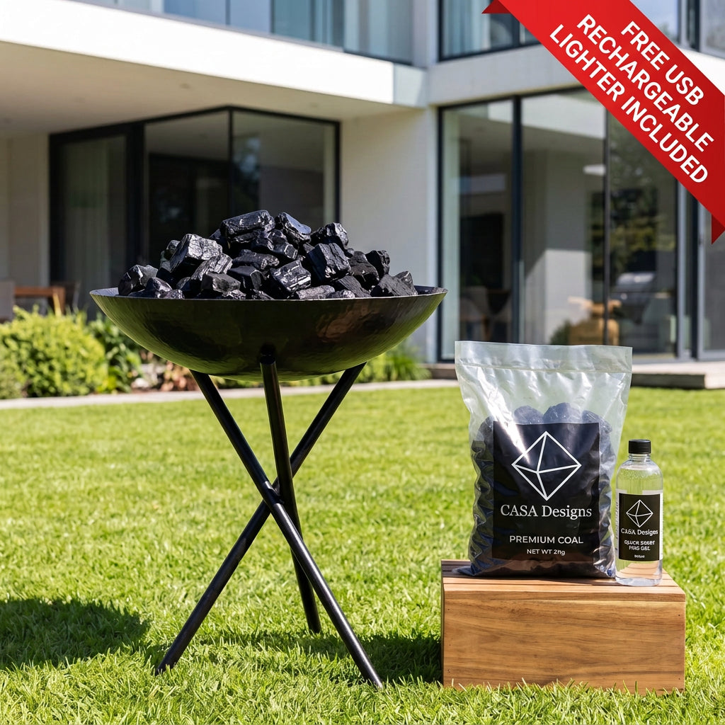 A black metal fire pit bowl filled with charcoal on a stand, set on a green lawn in front of a modern house. A bag of Casa Designs premium charcoal and a small bottle sit on a wooden block beside it.