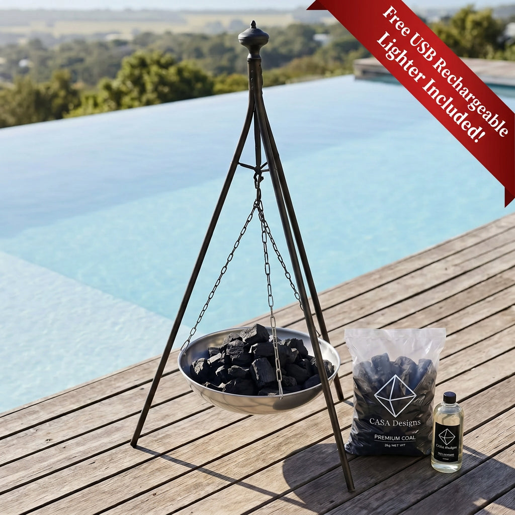 A metal tripod-style hanging fire pit bowl filled with charcoal sits on a wooden deck beside a clear bag of Casa Devissot premium charcoal and a small bottle, with an infinity pool and outdoor landscape in the background.