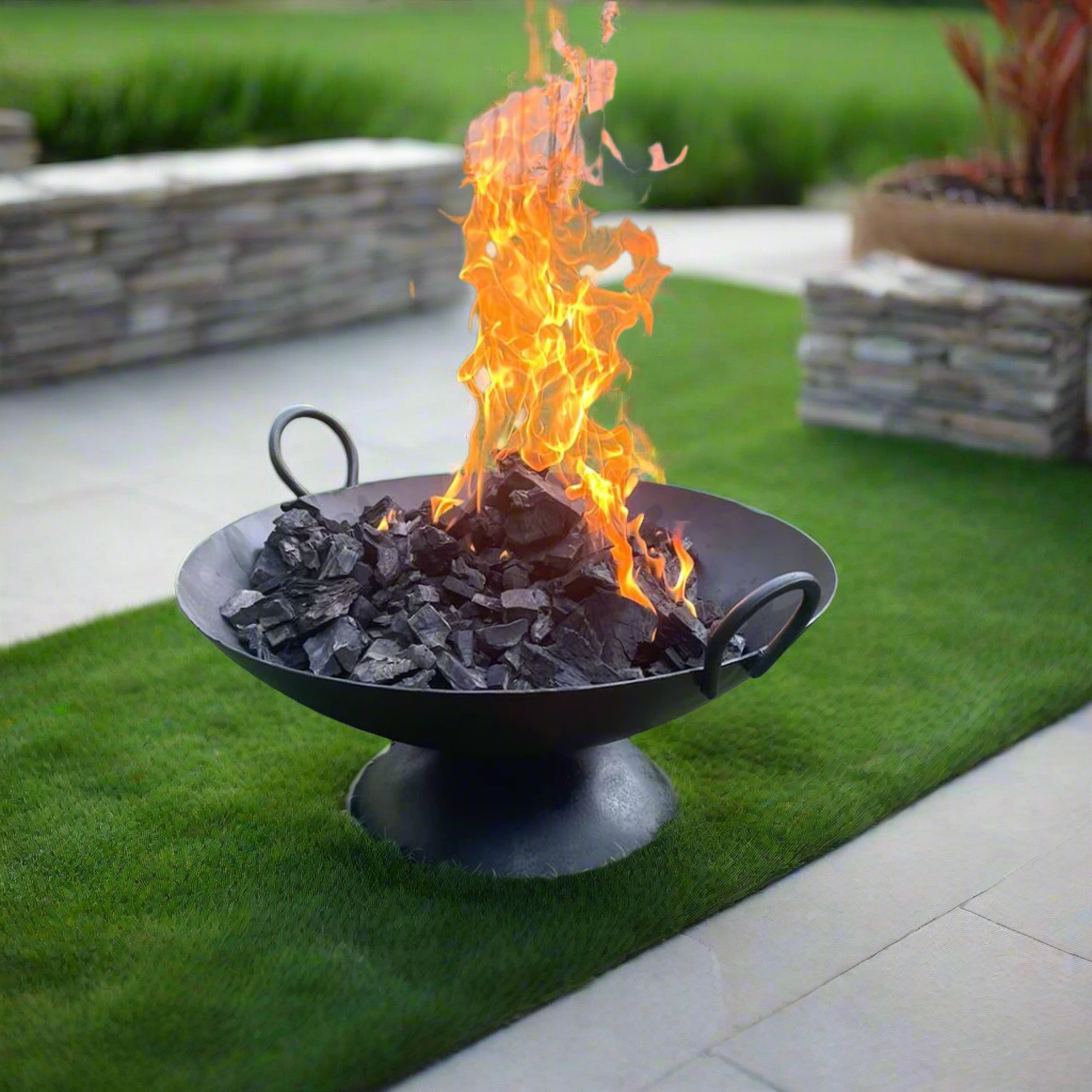 A large, round metal bonfire pit with wood inside,Igniting fire coming out from the wood, placed on a grassy area.